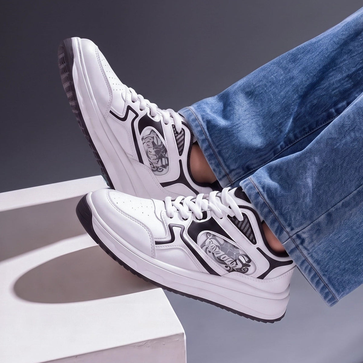 White low-top sneakers with black graphic details and white laces, shown on a model wearing blue jeans with legs crossed over a white geometric block against a dark gray background.